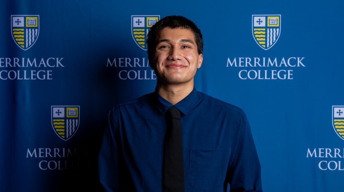 Smiling person in blue shirt and black tie posing before Merrimack College backdrop.