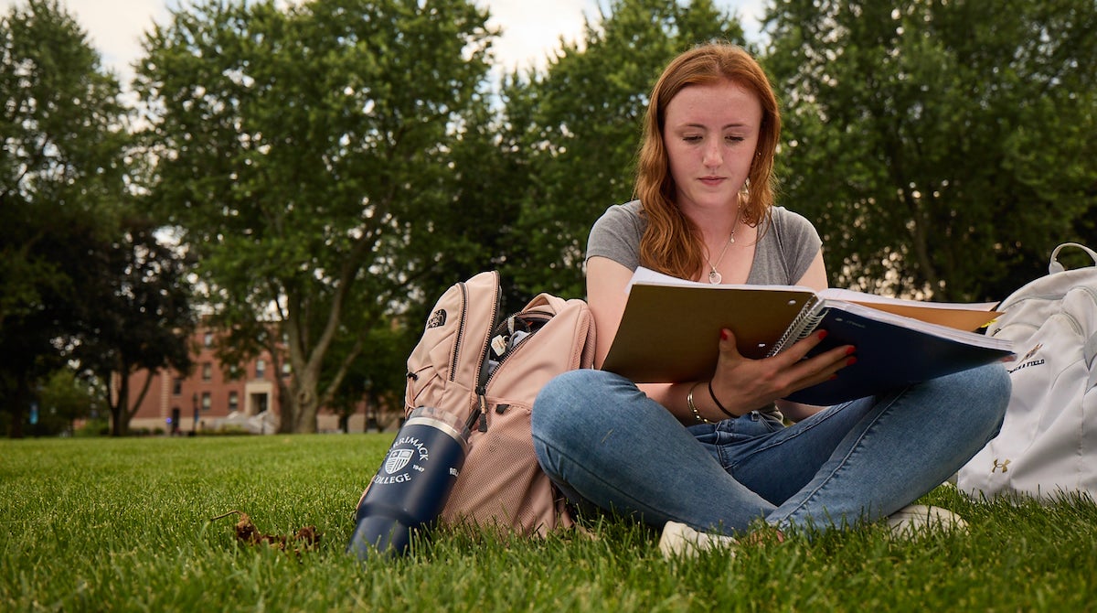 Photo of Allison Weed sitting on grass reading a book next to her backpack.