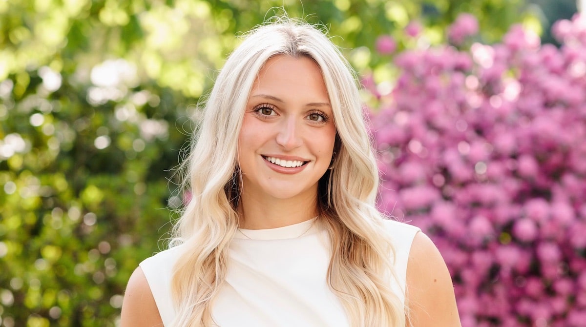 Smiling blonde woman stands outdoors, framed by green foliage and blooming pink flowers in sunlight.