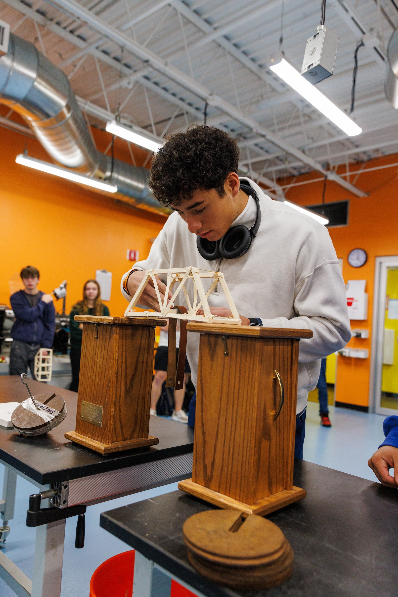 A student in a workshop carefully adjusts a small wooden truss bridge model placed between two testing supports while classmates observe in the background.