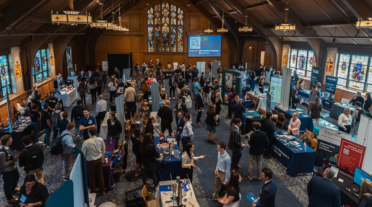 Crowded career fair inside a large hall with booths, attendees networking, and stained-glass windows visible.