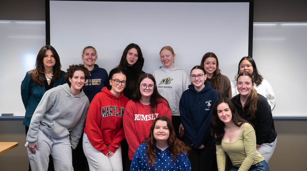A group of students and a teacher pose together in a classroom, smiling in front of a projector screen and whiteboards.