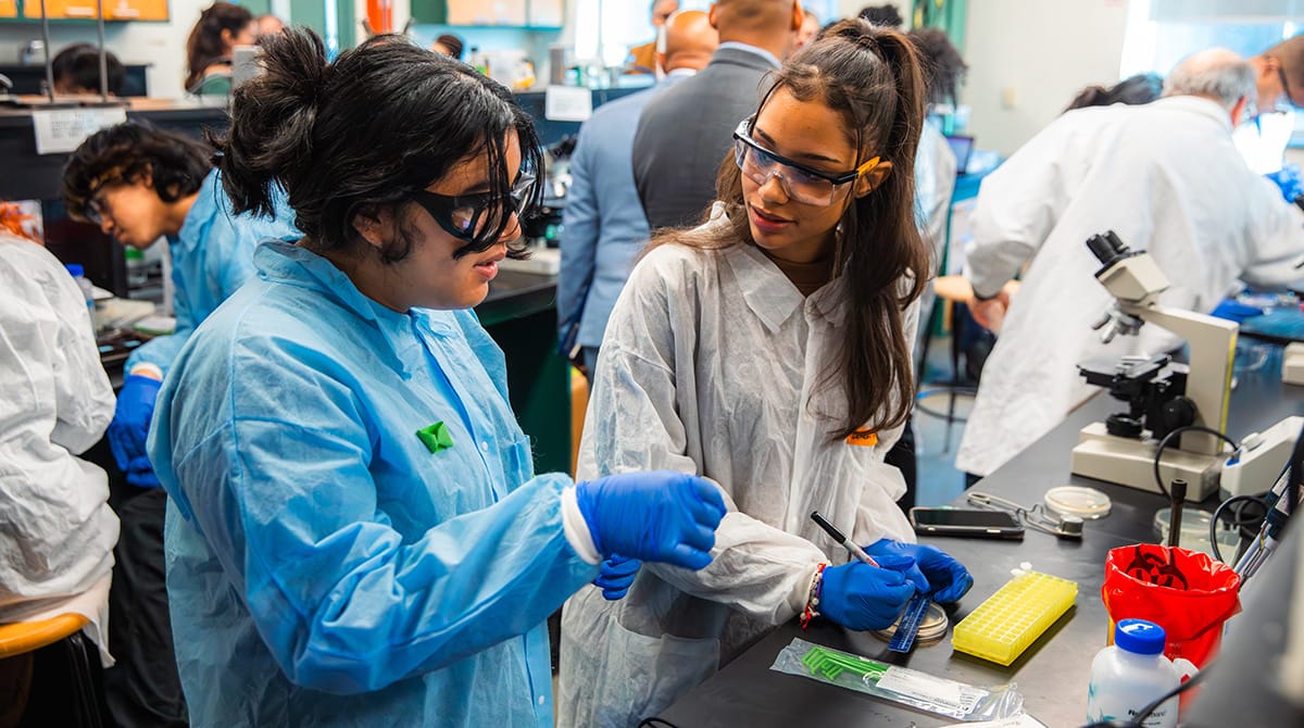 Photo of two students in scrubs and goggles working in a biology lab classroom.