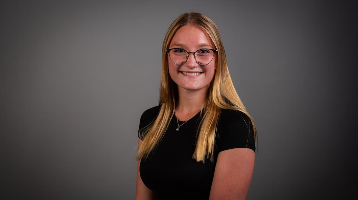 Smiling young woman with glasses and long blonde hair poses against a plain gray background.