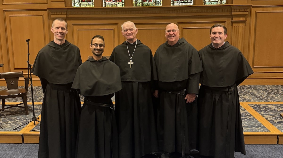 Five preists in black robes posing inside a church with stained glass windows.