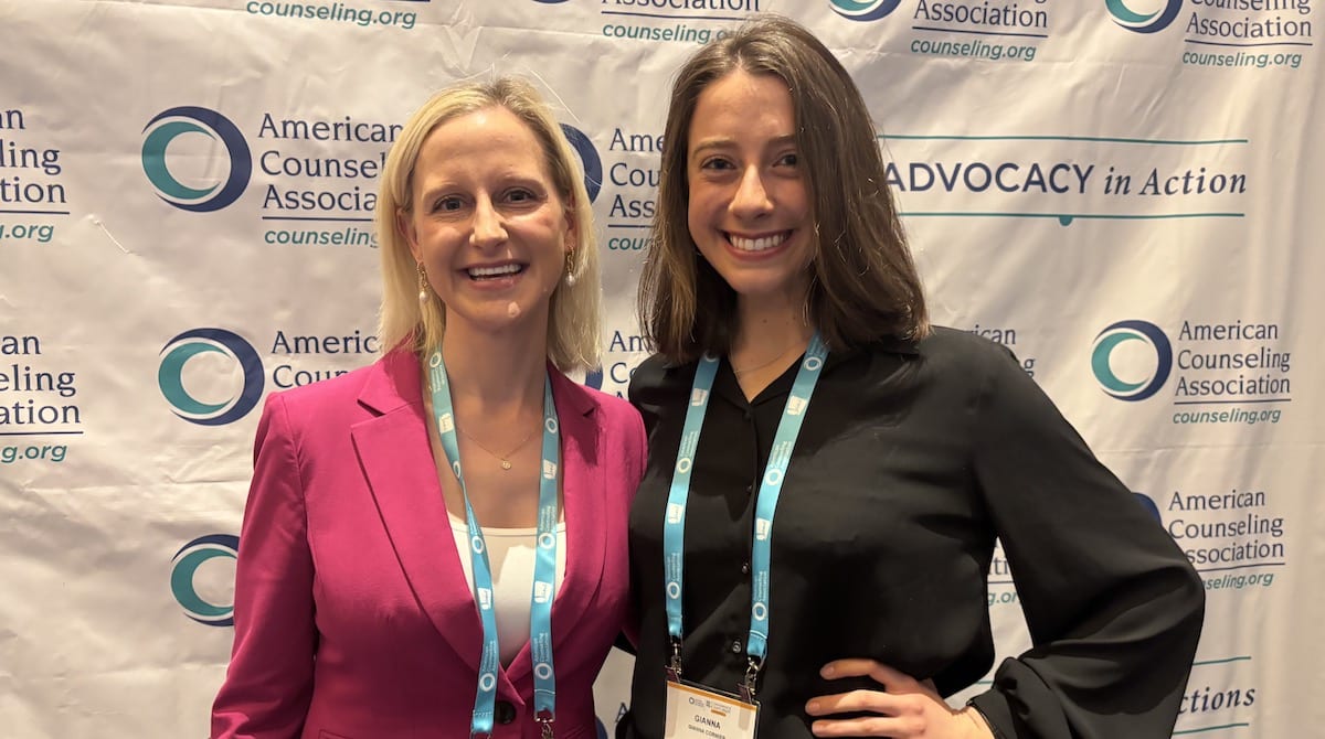 Two women smiling at American Counseling Association conference backdrop, wearing lanyards and name badges together.