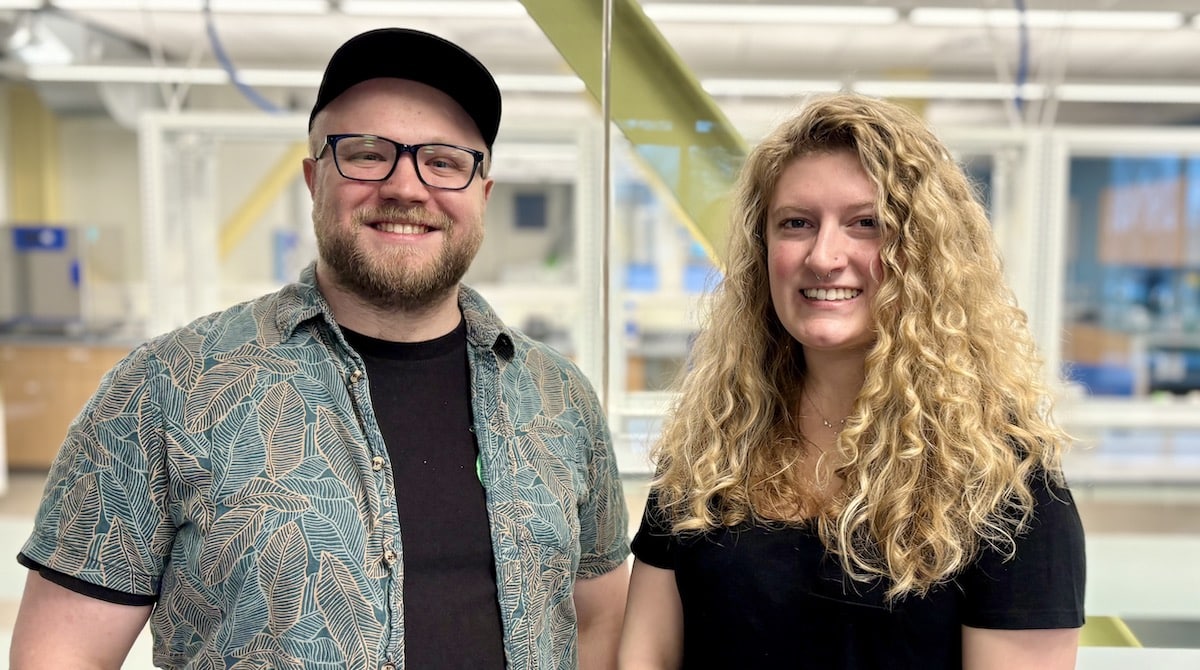 A man and woman smile warmly in a bright indoor setting. The man wears glasses, a cap, and a patterned shirt, while the woman has curly hair and a black top.
