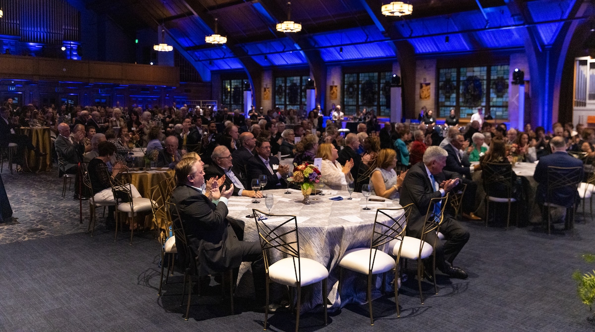 Guests seated at round tables in a warmly lit banquet hall applaud during a formal event.