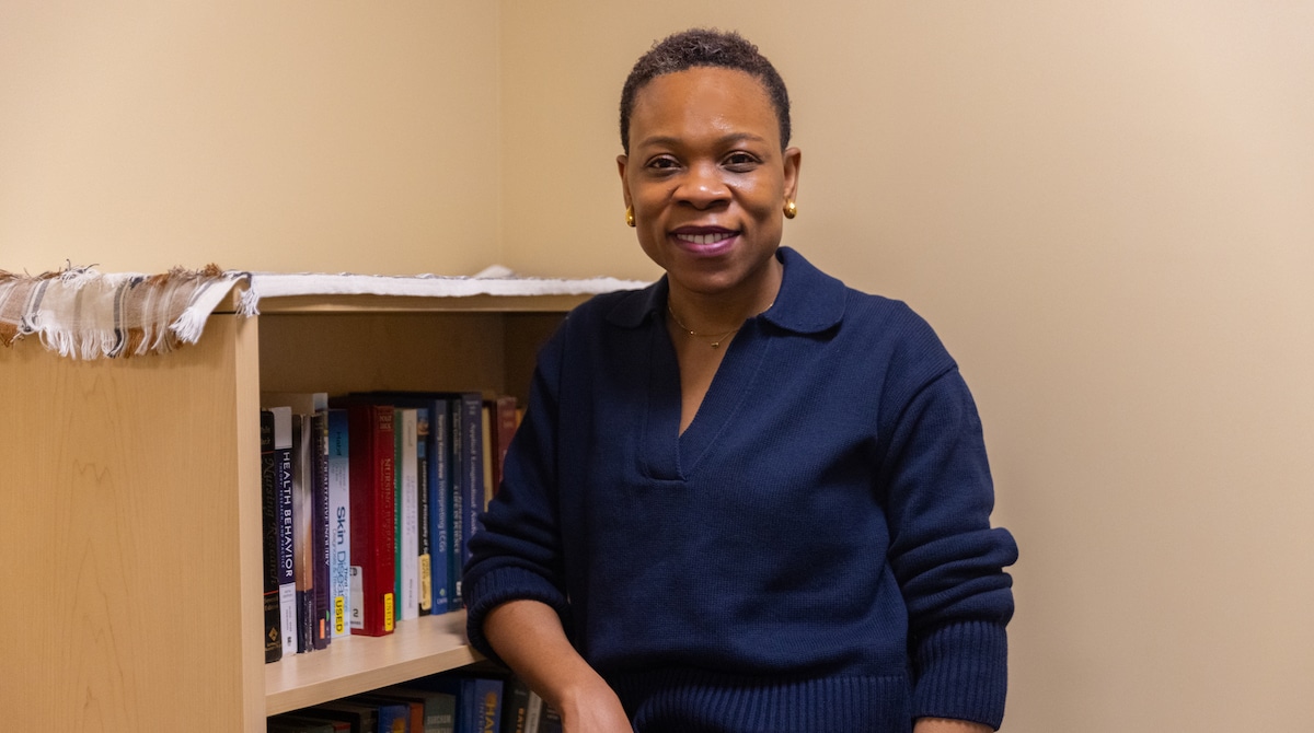 A woman with short hair and gold earrings smiles at the camera while standing beside a bookshelf filled with books in a warmly lit office setting.