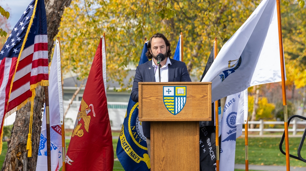A speaker addresses an outdoor audience at a podium surrounded by military and national flags.