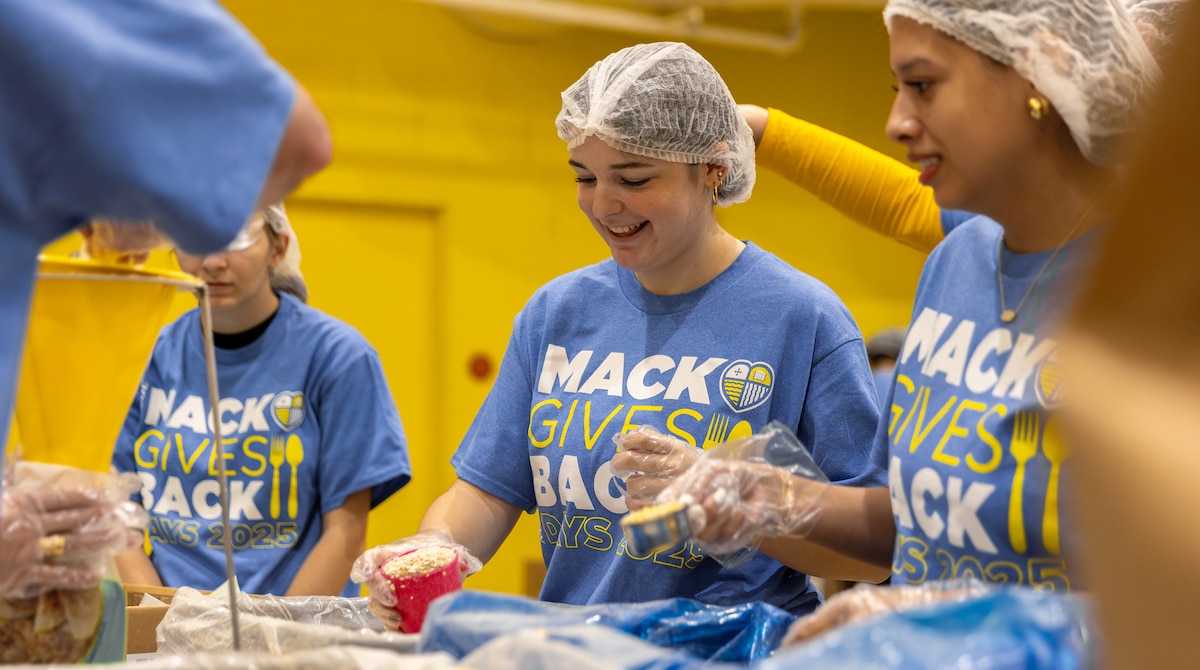 Smiling volunteers in hairnets and blue “Mack Gives Back” shirts prepare food together at a service event.