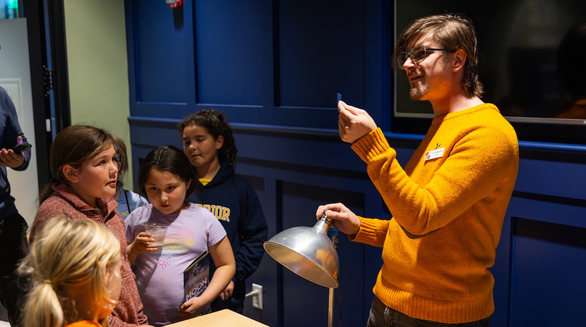 Man in orange sweater demonstrates with light and small object to group of attentive children indoors.