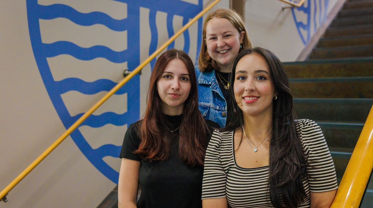 Three smiling young women pose together on a staircase with a Merrimack College logo mural in the background.