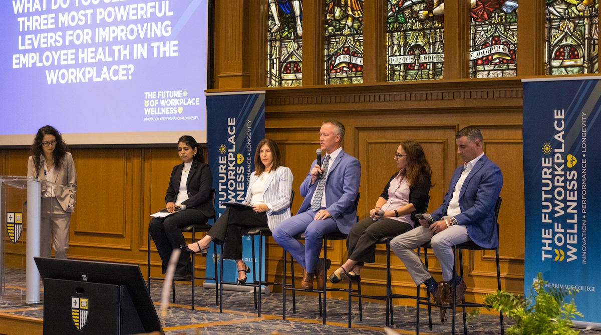 Panelists seated on stage discuss employee health and wellness, with moderator and presentation screen displayed.