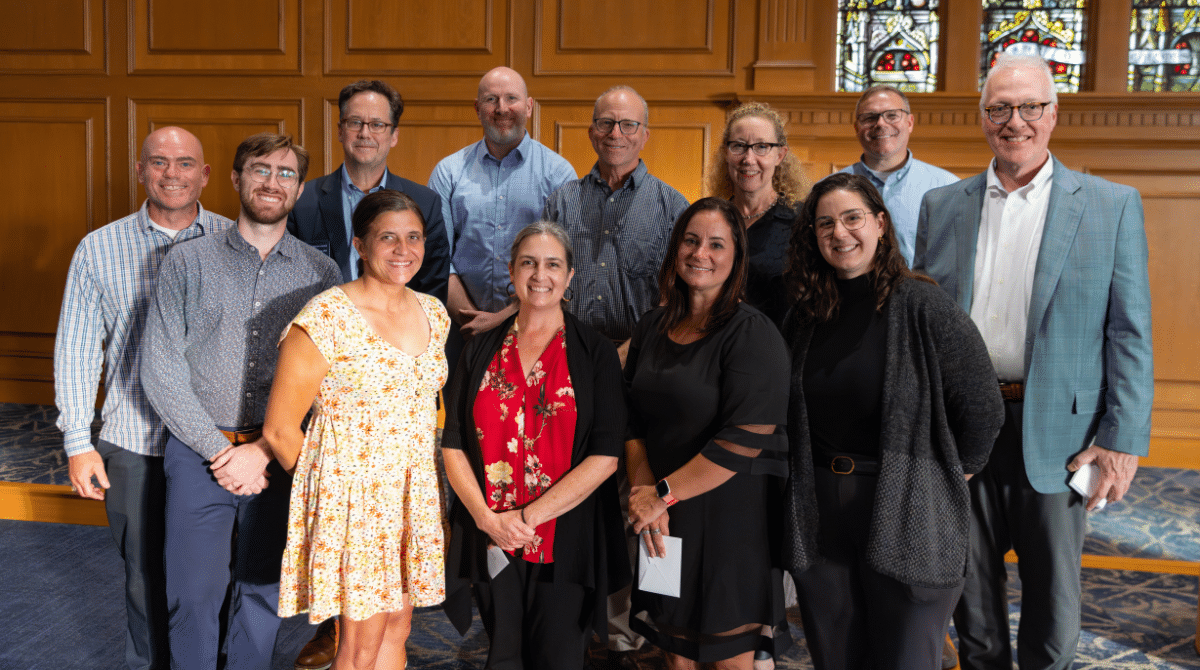 Group of smiling adults pose together indoors in front of wood paneling and stained glass windows.