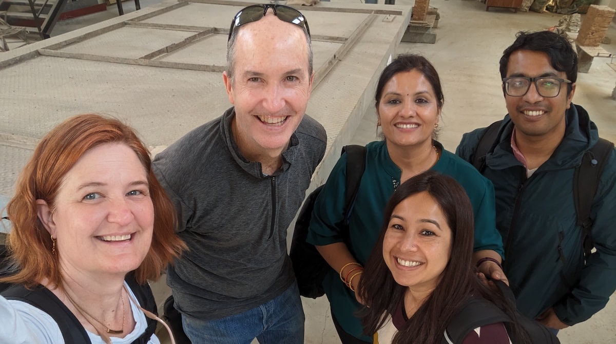 Five smiling adults taking a group selfie inside an archaeological site with stone foundations visible behind them.