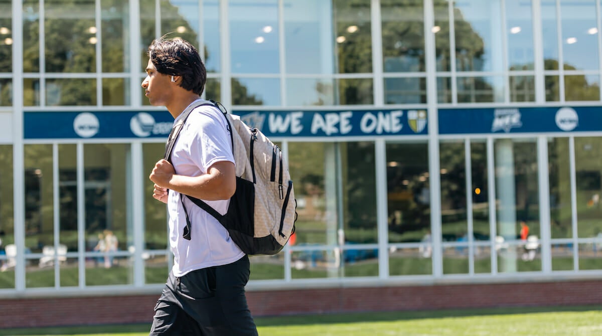 Photo of a Merrimack Student walking past a sign that reads, "We Are One."