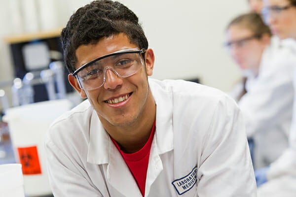 Student smiling in lab wearing lab coat and lab glasses