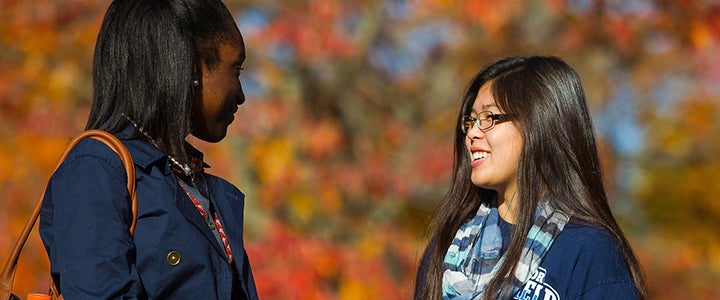 Two students talking to each other outside in front of fall foliage