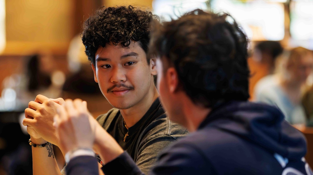 Two men sit across from each other in a warmly lit room, one in sharp focus with curly hair and a slight mustache, hands clasped as he listens, while the other is blurred in the foreground facing him.