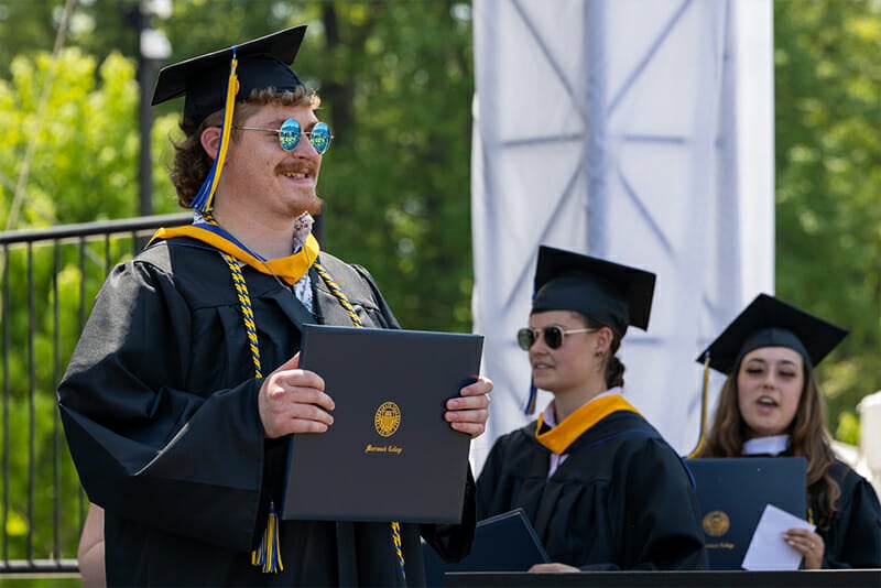 Student wearing round sunglasses holding up Merrimack diploma at Commencement. 