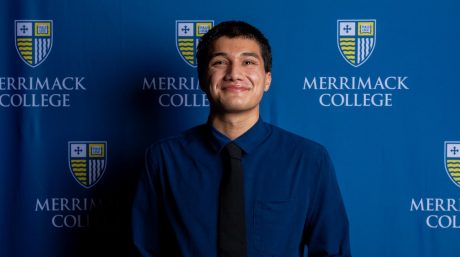 Smiling person in blue shirt and black tie posing before Merrimack College backdrop.