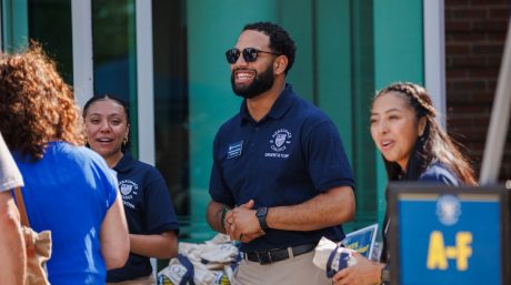 Three orientation leaders in navy shirts smile and engage with new students outside Merrimack College.