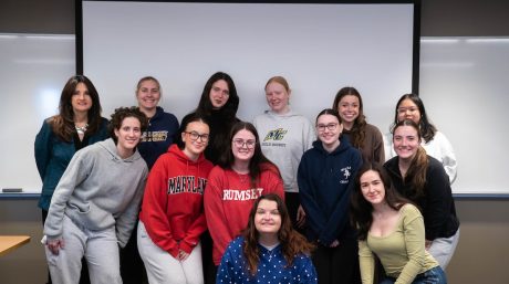 A group of students and a teacher pose together in a classroom, smiling in front of a projector screen and whiteboards.