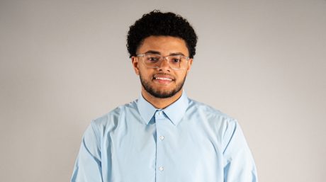 Person wearing glasses and light blue button-down shirt, standing against a neutral studio background.