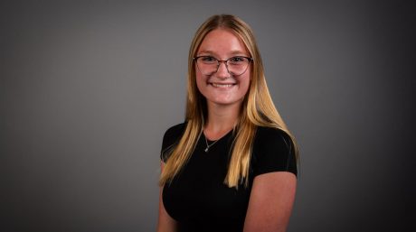 Smiling young woman with glasses and long blonde hair poses against a plain gray background.