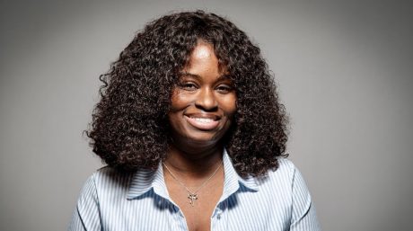 Smiling woman with curly hair wearing striped shirt and necklace against neutral gray studio background.