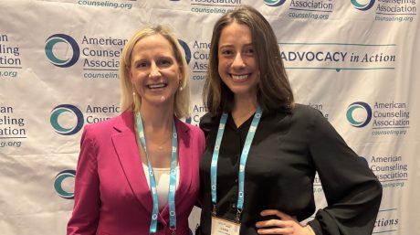 Two women smiling at American Counseling Association conference backdrop, wearing lanyards and name badges together.
