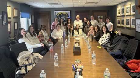 Group of about 15 people seated around a long conference table, smiling at the camera in a warmly lit meeting room with framed artwork and a sports jersey on the wall; water bottles, notebooks, and drinks are spread across the table.