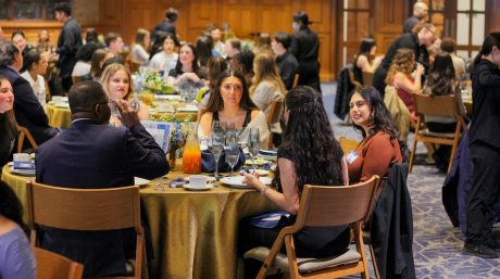 People seated at round tables during a formal banquet, talking and dining in a large wood-paneled event hall.