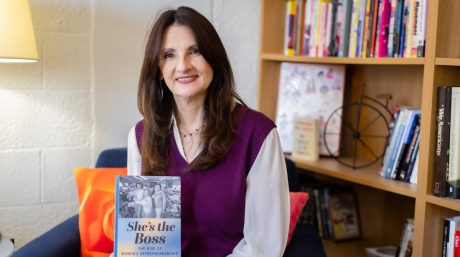 Woman smiling in office, holding book titled “She’s the Boss: The Rise of Women’s Entrepreneurship,” bookshelf behind her.