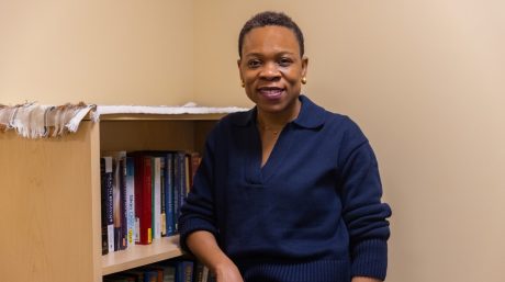 A woman with short hair and gold earrings smiles at the camera while standing beside a bookshelf filled with books in a warmly lit office setting.