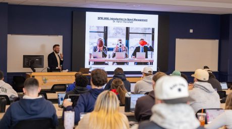 College professor lectures in a packed classroom while a sports management presentation displays on a large screen.