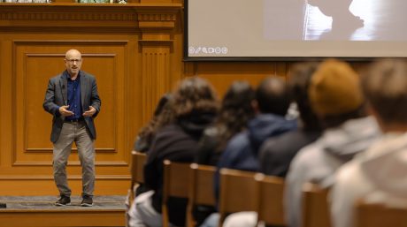 A speaker presents to an audience in a wood-paneled lecture hall with a projected image.