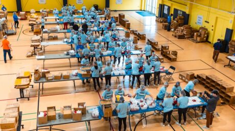 Groups of volunteers circle around tables packing non-perishable meals