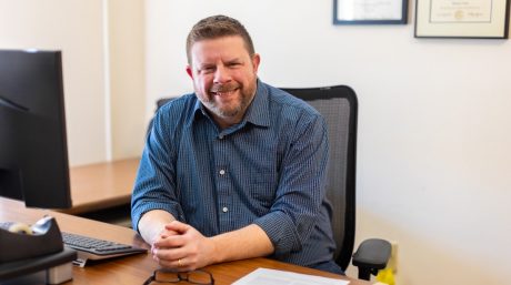 Man in blue checkered shirt smiling at desk in office setting, hands clasped near keyboard and glasses.