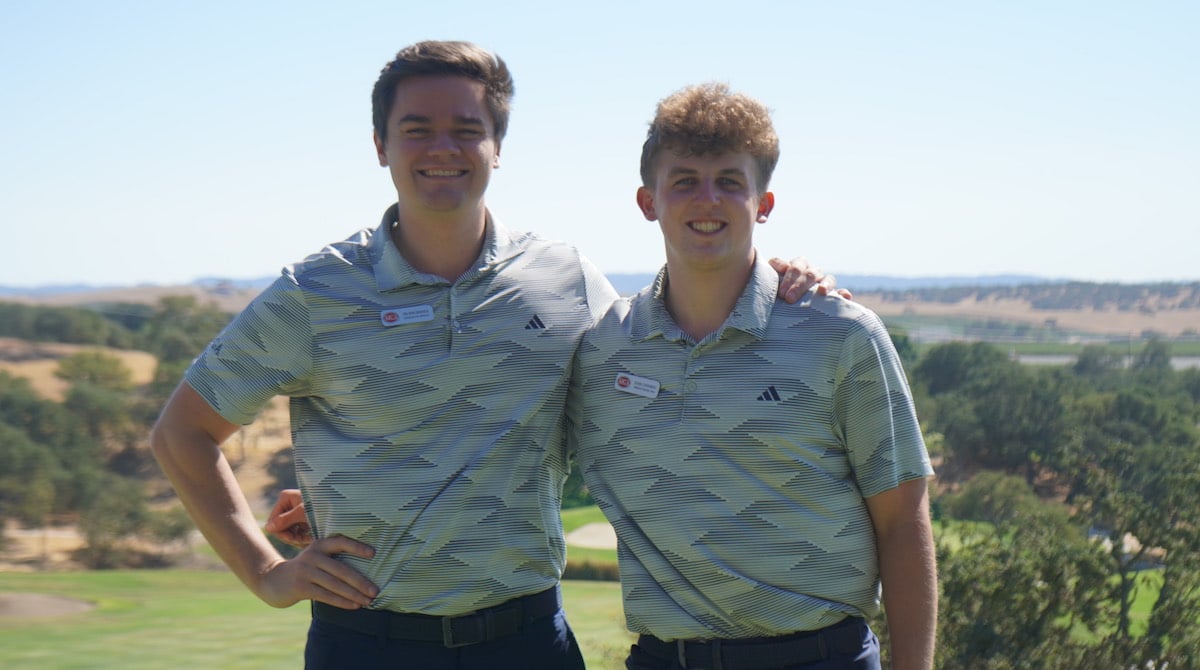 Two smiling men in matching polo shirts stand together outdoors on a sunny golf course.